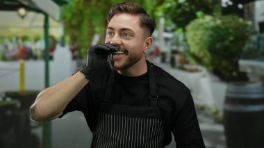 Sakallı ve önlüklü genç beyaz adam, bir şehir caddesinde garsonluk yapıyor, açık şehir ortamında eldivenle gülümsüyor..