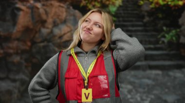 Açık havada gülümseyen, gönüllü kırmızı bir rozet takan, sarı saçlı arka planda merdivenleri çıkan, gençlik enerjisi ve toplum nişanı sergileyen bir kadın..