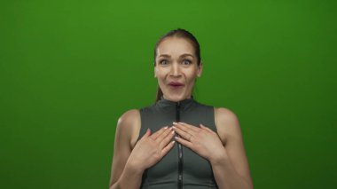 Stüdyoda kolsuz yelek giyen, geniş gözlü ve ağzı açık yeşil bir duvara parmak göğsü gösteren bir kadın..