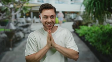 Sakallı genç bir adam şehir caddesinde şaşkın bir şekilde beliriyor, elleri açık yeşil bir arka planda, neşeli bir hareketle gözlerini örtüyor..