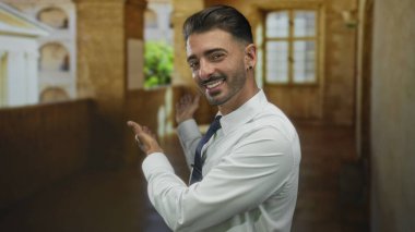 Beyaz tişörtlü genç İspanyol adam güneşli bina koridorunu işaret ediyor..