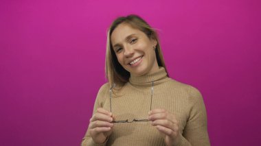 Boğazlı kazakla gülümseyen, canlı pembe arka planda duran, neşeli ifadeli gözlükleri ayarlayan ve neşeli havayı yansıtan rahat duruşu olan kadın..