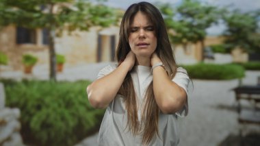 Şehir sokaklarında gezen Latin kökenli genç bir kadın yeşillik ve sıcak taş binalar arasında stresli bir şekilde boynunu tutuyor. Şehir yorgunluğu ve güzelliği çağrıştırıyor..