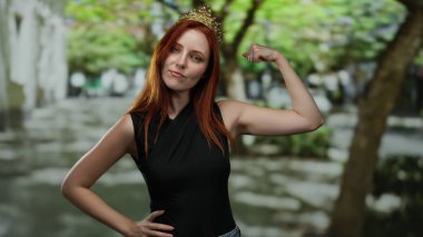 Kızıl saçlı ve taç takan bir kadın şehir sokaklarında esniyor, kendine güveni ve güzelliği açık şehir arka planında canlı yeşillikler sergiliyor..