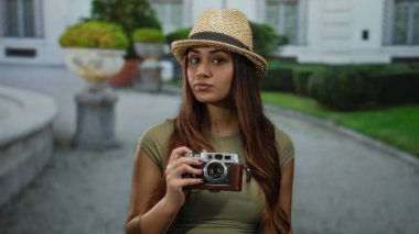 Sokakta elinde kamera olan bir kadın. Şapka takıyor ve binanın yakınına bakıyor. Seyahat ve merak uyandırıyor..