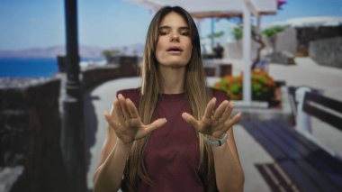 Uzun saçlı bir kadın, açık bir açık havada, arka planda canlı çiçekler ve güneşli bir sahil kenarında elleri ile sakince el kol hareketleri yapar..