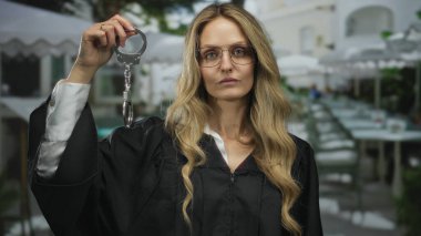 Kadın yargıç bir restoranın terasında kelepçe tutuyor, açık havada üniforma giyiyor, sarı saç, resmi ifade, sokak dekorasyonu, güneşli bir gün, kentsel çevre, adalet teması.