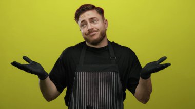 Önlük ve eldiven takan sakallı genç adam parlak sarı arka planda duruyor ve neşeli bir ifadeyle omuz silkiyor..