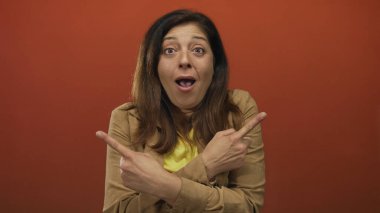 Orta yaşlı bir kadın, el hareketlerini vurgulayan turuncu bir stüdyo odasında yan yan bakarken parmağıyla objeyi işaret ediyor..