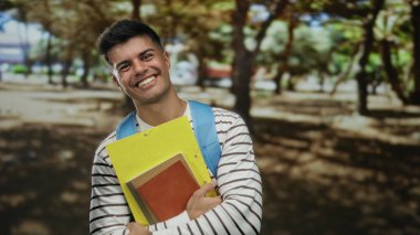 Genç adam parkta çizgili gömlek ve sırt çantası ile neşeli ifadeli defterler tutuyor parmaklarıyla zafer jestleri yapıyor..