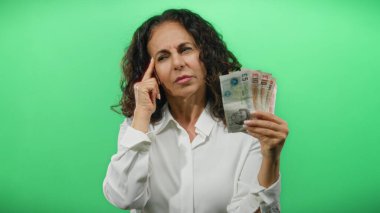 Orta yaşlı bir kadın yeşil bir geçmişe karşı İngiliz sterlini banknotları üzerinde kafa yoruyor, finansal karar verme ve tefekküre dalmayı somutlaştırıyor.