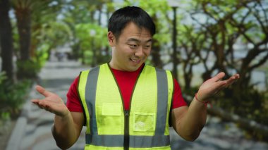 Kentsel sokak ortamında gülümseyen bir adam yansıtıcı yelek giyiyor, elleri açık genç bir adam yeşil ağaçların altında..