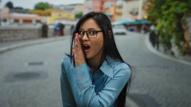 Mavi gömlekli genç bir kadın canlı bir şehir caddesinde fısıldıyor, renkli binaların arka planına sahip samimi, neşeli bir açık hava anını yakalıyor..