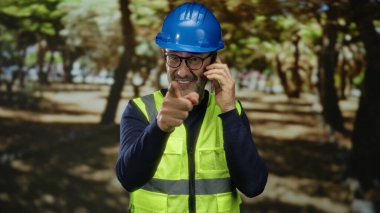 İnşaat malzemesi giymiş olgun bir adam açık hava parkında jestlerle telefonda konuşuyor..