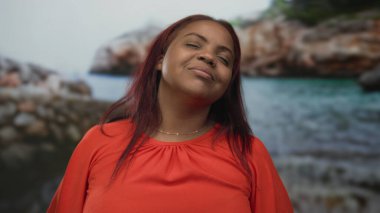 Turuncu giysili kadın, güneş ışığı altında kayalık sahil manzarasında gözleri kapalı, başını arkaya yaslıyor..