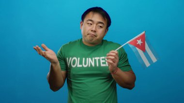 Yeşil gömlekli genç adam mavi arka planda Küba bayrağı tutuyor. Gönüllü desteğini düşünceli bir ifadeyle gösteriyor..