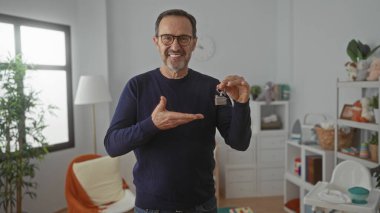 Gözlüklü olgun bir adam ev anahtarlarını kucağında bebek dekoruyla sıcak ve mutlu bir şekilde yatak odasında tutuyor..