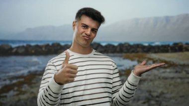 Sahilde duran genç adam başparmaklarını kaldırarak ve açık bir el göstererek arka planda dağlar ve bulutlu gökyüzü altında dalgalar olan çizgili tişört giyerek kıyıda duruyor.