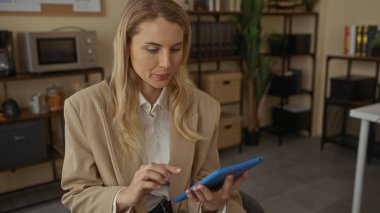 Ofis ortamında tablet kullanan ve arka planda raflar ve kahve makinesi olan bir kadın, meşgul ve kapalı bir profesyonel ortamda gülümsüyor..