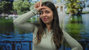 Yeşil yapraklar ve gölcüklerle çevrili güneşli bir parkta, açık havada el işareti yapan bir kadın. Parlak gökyüzünün altında neşe saçan bir enerji yayıyor..