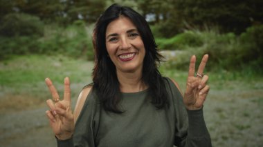 Orta yaşlı bir kadın yeşil park ortamında iki eliyle barış işareti yaparken geniş bir gülümseme ve zeytin gömleği giyiyor..