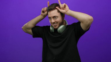 Sakallı, kulaklık takan genç adam mor duvara karşı şakacı bir jest yapıyor. İzole edilmiş, dışlanmış, dışlanmış, dışa dönük, dışavurumcu, neşeli ve modern..