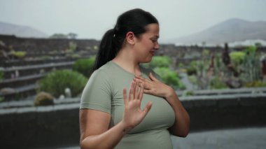 Kıvrımlı vücutlu genç bir kadın park ortamında reddedici bir jest yapıyor. Bir eli göğsünde, açık hava yeşillikleriyle çevrili bir şekilde hoşnutsuzluğunu dile getiriyor..