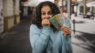 Bir kadın endişeyle bir şehir caddesinde elinde banknotlarla mavi bir kazak giyiyor, mali kaygıları ve duyguları temsil ediyor..