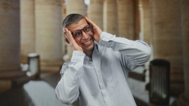 Gözlüklü ve neşeli ifadeli bir adam dışarıda durur, neşeli bir şekilde yüzünü kapatır ve sütunların altında bir şehirde gülümser, şehir ortamında mizah ve duygu gösterisi yapar..