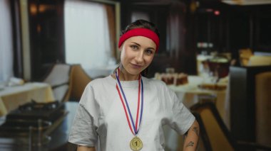 Madalyon takan bir kadın restoranda kendine güvenerek ayakta duruyor. Kırmızı bir saç bandıyla şık bir yemek geçmişine karşı günlük giysiler içinde atletik başarılar sergiliyor..