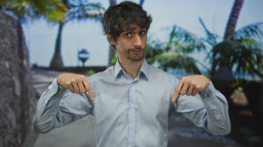 Mavi gömlekli genç adam, yeşil orman ortamında işaret parmaklarıyla göğsünü işaret ediyor..