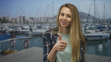 Arka planda teknelerle marinaya gülümseyen, sıradan kıyafetler giyen ve güneşli bir günde baş parmak işareti yapan genç bir kadın..