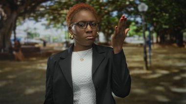 Siyah ceketli ve beyaz elbiseli kadın parkta sık yapraklı ağaçların altında el ele tutuşuyor..