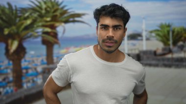 Beyaz gömlekli genç İspanyol adam sahilde kalçasını dinlendirirken kollarını ve başını eğiyor; dinginlik.