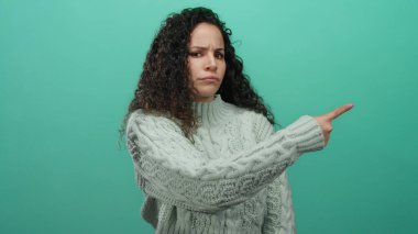Süveter giymiş genç bir İspanyol kadın sert bir ifadeyle işaret ediyor. Etkileyici iletişim temaları için parlak yeşil bir arka planda izole edilmiş..