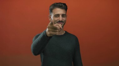 Yeşil kazaklı genç İspanyol adam stüdyo ortamında kamerayı işaret ediyor; güven motivasyonu başarı hırsı.