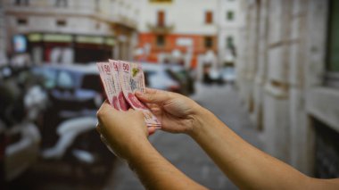 Bir şehir caddesinde Meksika pezosu tutan adam, açık hava mali işlemleri ve görünür park edilmiş arabaların olduğu bir şehir ortamına yatırım göstermektedir..