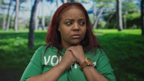 Женщина в волонтерской рубашке сжимает руки за грудь в лесу в окружении деревьев и зелени; тревога.