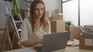 Yeni ev binasındaki ahşap masadaki paketsiz karton kutuların arasında dizüstü bilgisayarda yazan kadın; odaklanma planlaması.