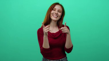 Neşeli bir gülümsemeyle kamerayı gösteren bir kadın, kırmızı saçlı, kırmızı bir kazak giyen, parlak yeşil duvarlı bir arka plan.