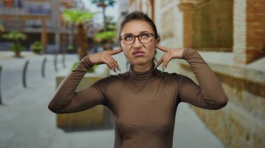 Hayal kırıklığına uğramış bir ifadeyle gözlük takan genç bir kadın şehir sokaklarında kulaklarını tıkıyor.