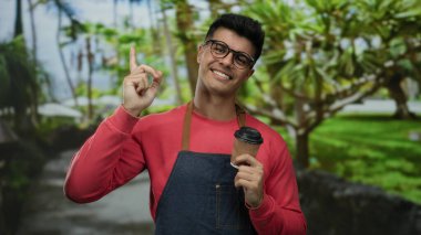 Gözlüklü, kırmızı ve önlüklü, elinde kahve fincanı tutan genç adam, gülümsüyor ve yemyeşil bir arka planda yukarı doğru bakıyor..