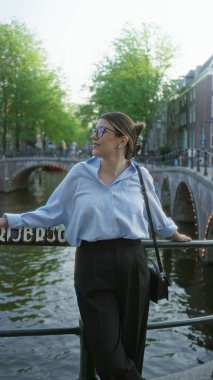 Amsterdam 'da gülümseyen, köprüler ve yeşilliklerle dolu bir kanalda duran mavi bluz ve gözlük takan, gün ışığında şehir manzarasını gösteren bir kadın..