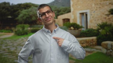 Beyaz bir adam açık hava parkında göğsüne gözlük takıyor. Arka planda kırsal bir bina ve yeşillik var. Güvenini ve rahatlığını aktarıyor.