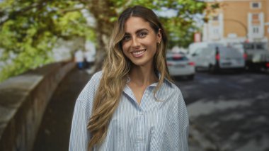 Genç sarışın kadın yüzü, çizgili bluz içinde dalgalı saçlarıyla gülümsüyor yapraklı güneş ışığı kaldırımında; keyif güveni.