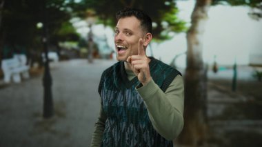 Şehir sokaklarında gülümseyen ve kameraya bakan genç adam şehir ağaçlarının altında neşeli yüz ifadesini sergiliyor..