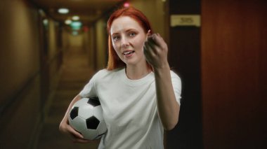 Otel koridorunda elinde futbol topu tutan, kendine güvenen bir kadın..
