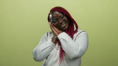 Afro-Amerikalı bir kadın sarı arka plan duvarında uyuyor gözleri kapalı ve elleri beyaz kıyafetler ve pembe örgülerle birlikte..