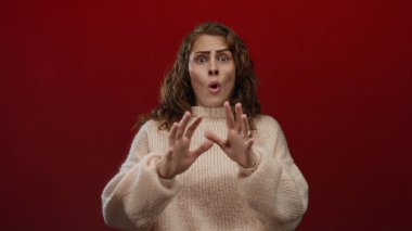 Kadın, canlı kırmızı bir arkaplan karşısında şaşırmış, rahat bir süveter giyiyor, duygularını ifade eden gözler ve ellerle yakalıyor, yaratıcı projeler için tamamen izole edilmiş..