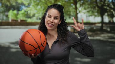 Genç İspanyol kadın elinde basketbol topuyla açık hava parkında jest yapıyor. Ağaçlarla çevrili güneşli gökyüzünün altında enerji ve coşku gösterisi yapıyor..